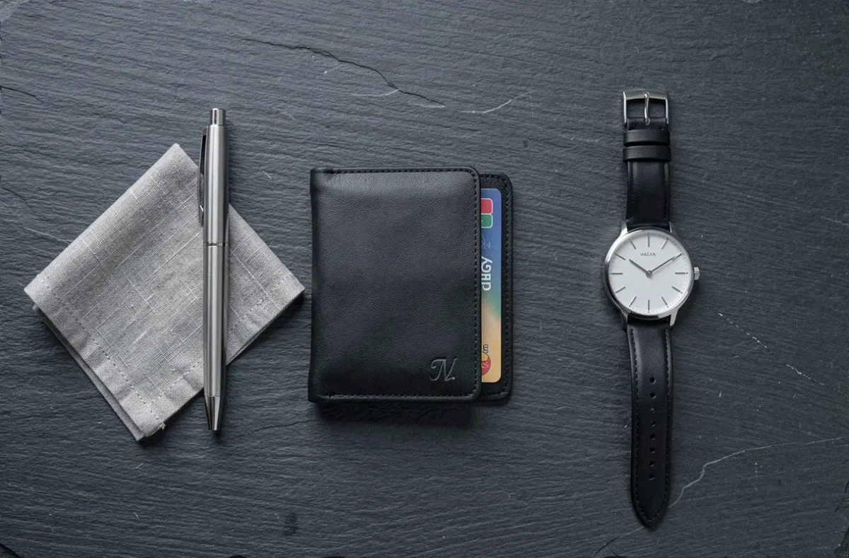 Flat lay minimaliste avec portefeuille cuir noir personnalisé initiales gravées stylo et montre sur ardoise sombre